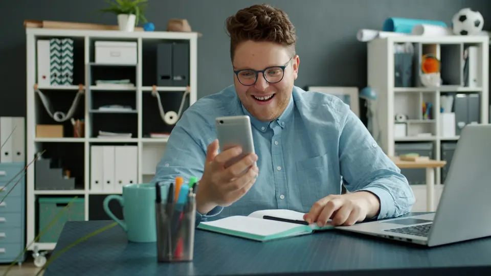 Mitarbeiter lacht am Schreibtisch beim Blick aufs Smartphone